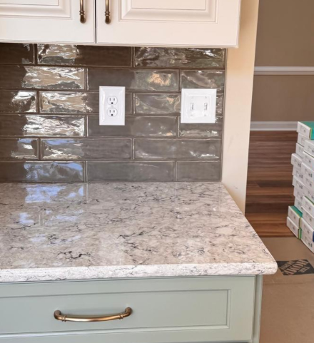 Kitchen countertop with gray backsplash tiles, white cabinets, and electrical outlets above a speckled stone counter