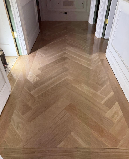Narrow hallway with light wood herringbone floor and white doors leading to a room.