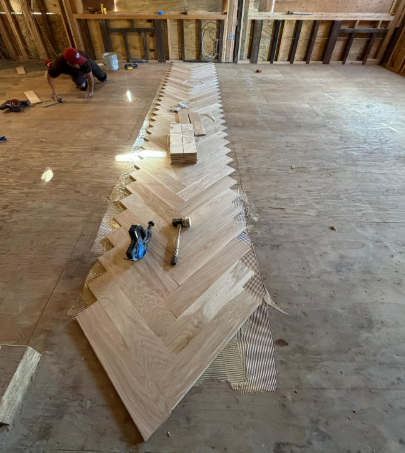 Herringbone wood flooring being installed on a subfloor in an unfinished room, with a worker kneeling nearby