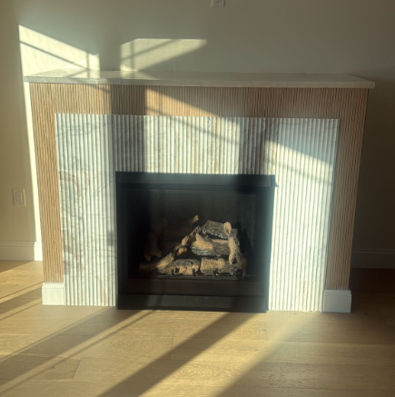 Sunlit living room fireplace with a black firebox, wood-paneled surround, and patterned light on the floor