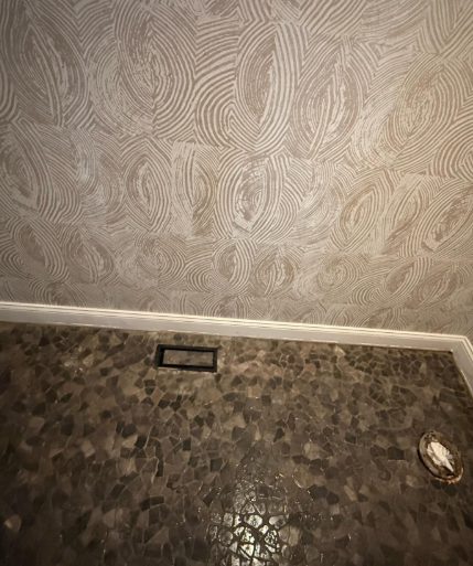 Empty corner with patterned beige wallpaper, white baseboard, and dark speckled floor with a floor vent.