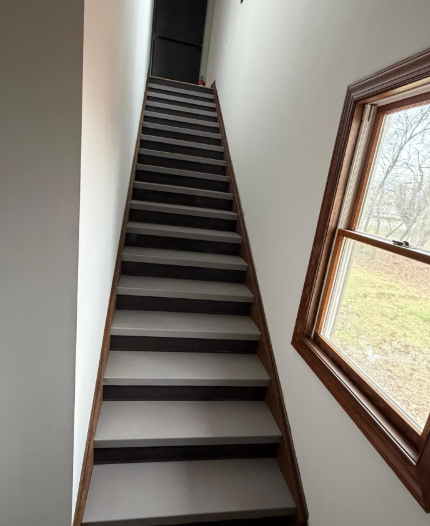 Steep indoor staircase with dark treads, white walls, and a window beside it