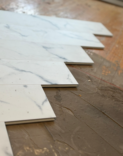 Stacked white marble-look floor tiles on a brown wooden floor, viewed from above