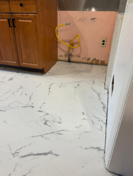 Kitchen with white marble-look floor, wood cabinets, and a pink wall with exposed plumbing.
