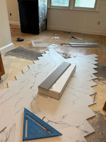 Flooring installation in progress with white marble-look tiles, tools, and a blue triangle on a hardwood floor