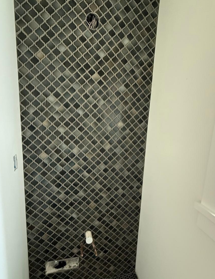 Tiled shower wall with gray mosaic tiles, shower fixtures, and a bottle on the floor