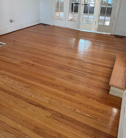 Empty room with polished hardwood floors and white walls, sunlight from glass doors.