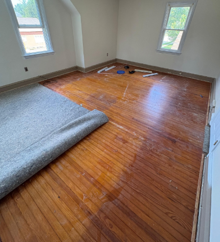 Empty room with hardwood floors, gray carpet roll, and two windows letting in daylight.