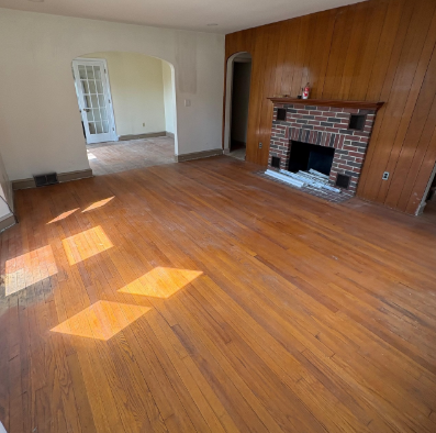 Empty living room with hardwood floors, brick fireplace, and sunlight streaming in from the windows.