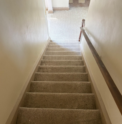 Carpeted staircase descending to a bright landing, with beige walls and a wooden handrail.