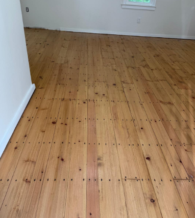 Empty room with unfinished wooden floor and white walls, lit by a small window.