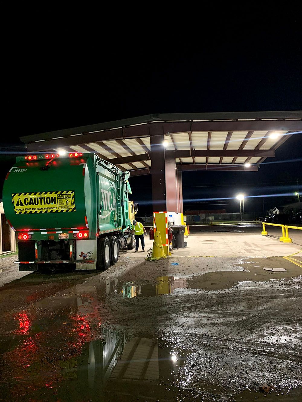 A green garbage truck is parked in a well-lighted parking lot