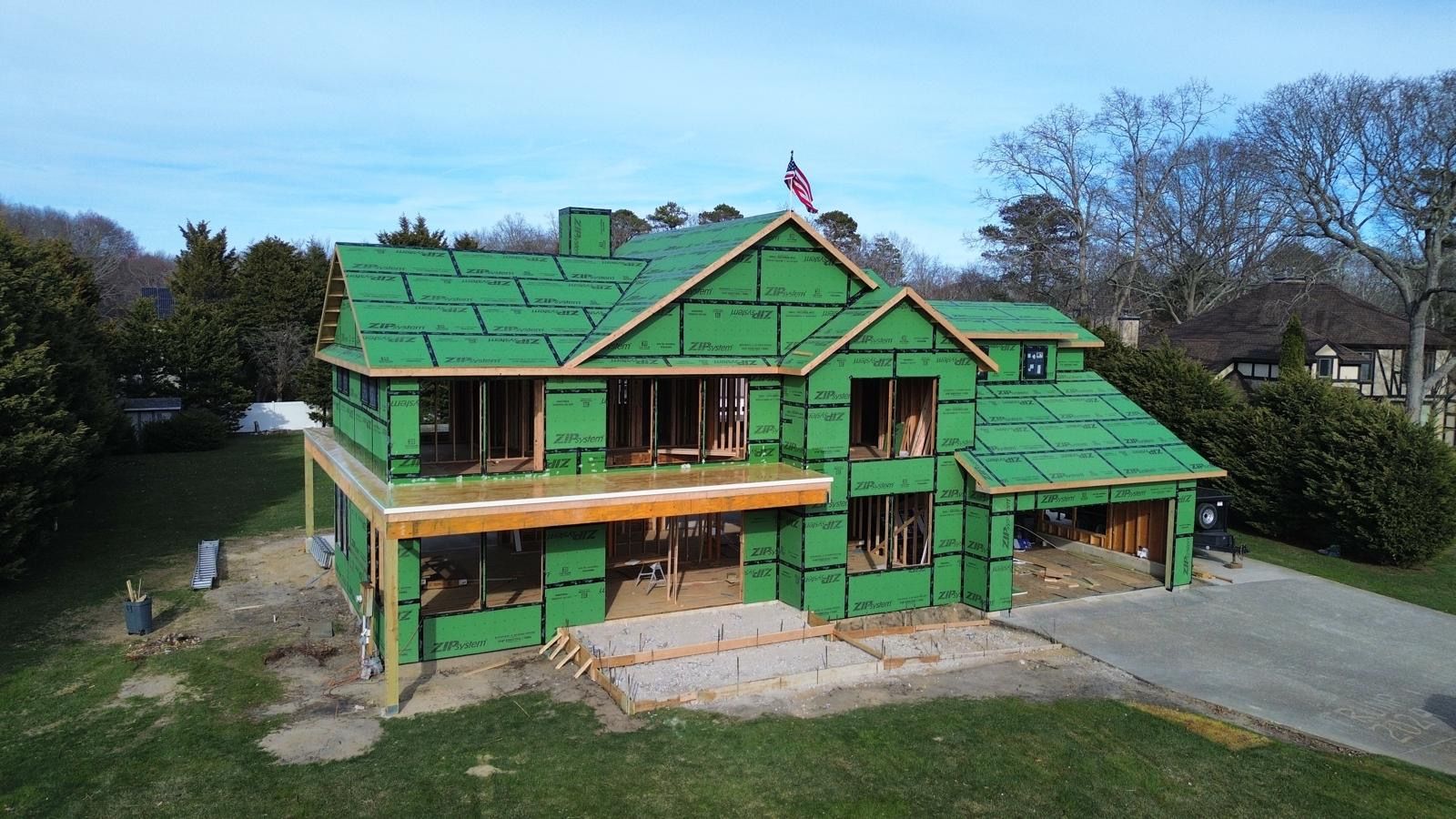 House under construction wrapped in green sheathing and scaffolding, set on a grassy lot with trees nearby