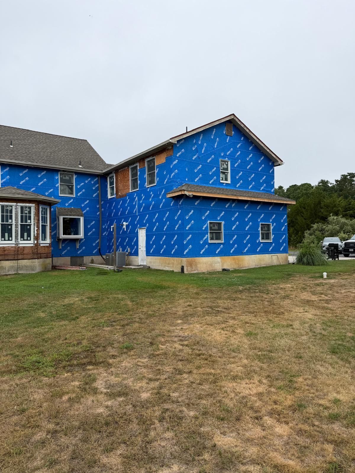 Blue house under construction with plywood wrap, grassy yard, and overcast sky.