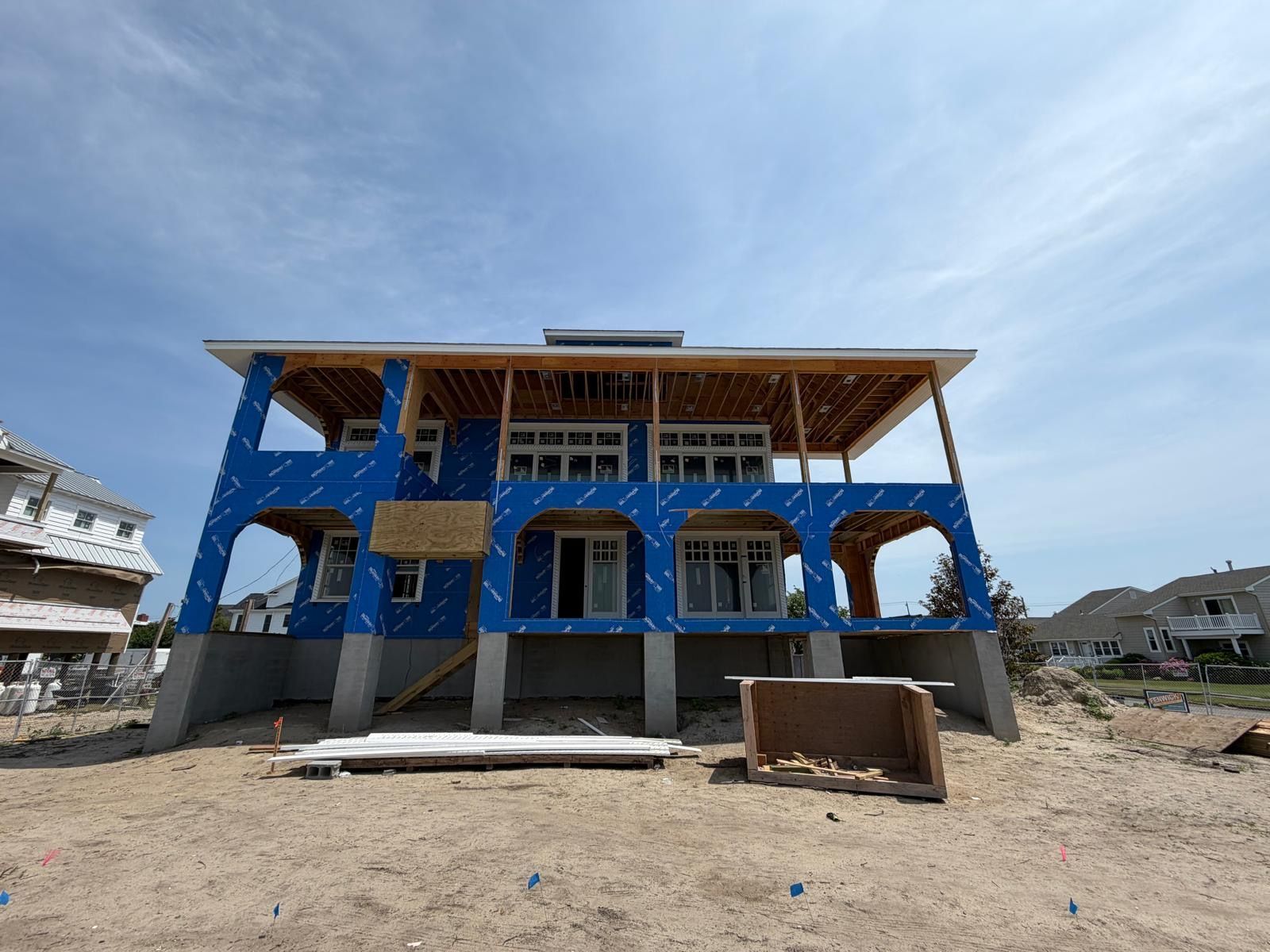 Blue coastal house under construction with wooden roof framing and sandy lot