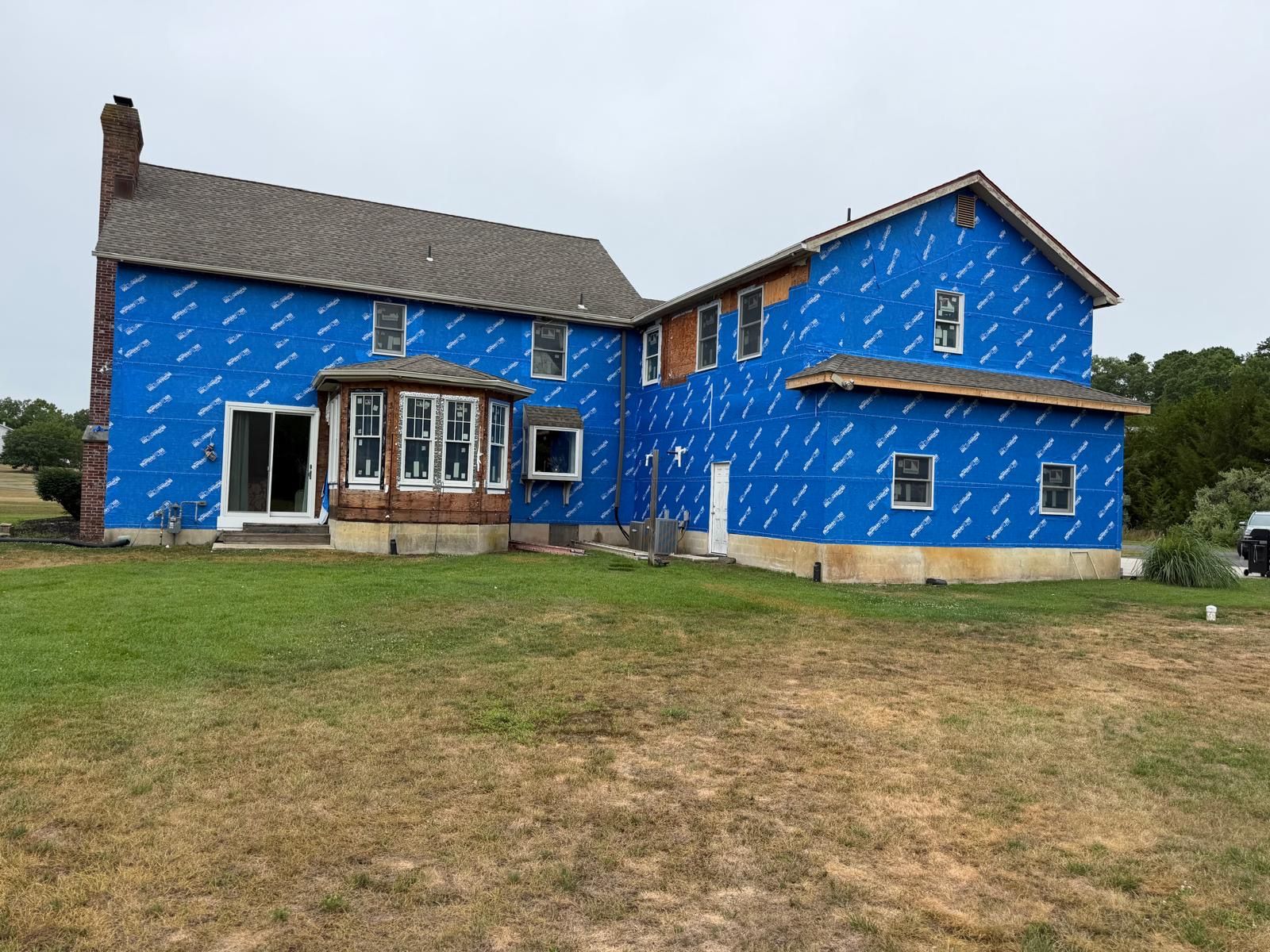 Partially built house wrapped in blue weather barrier, with a grassy yard and overcast sky