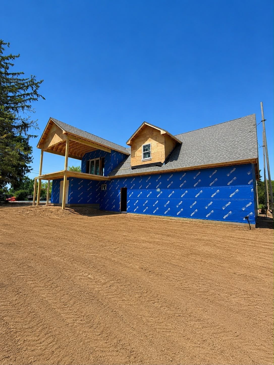 Blue house with tan roof and porch on a sloped dirt lot under a clear blue sky