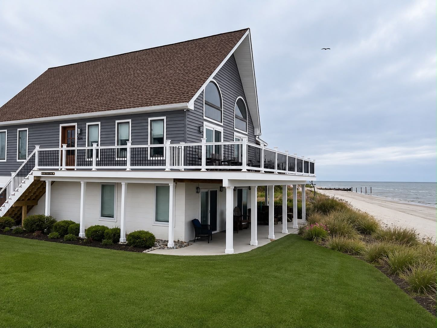 Gray beach house with white deck overlooking ocean and sandy shore under cloudy sky