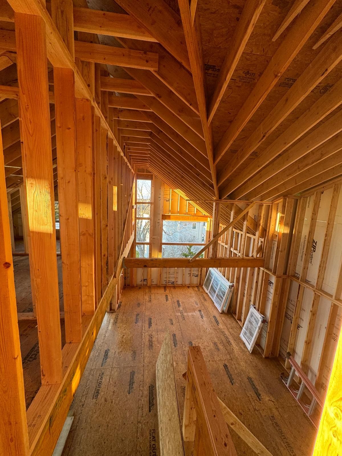 Narrow unfinished attic corridor with exposed wooden framing, angled rafters, and daylight through a window.