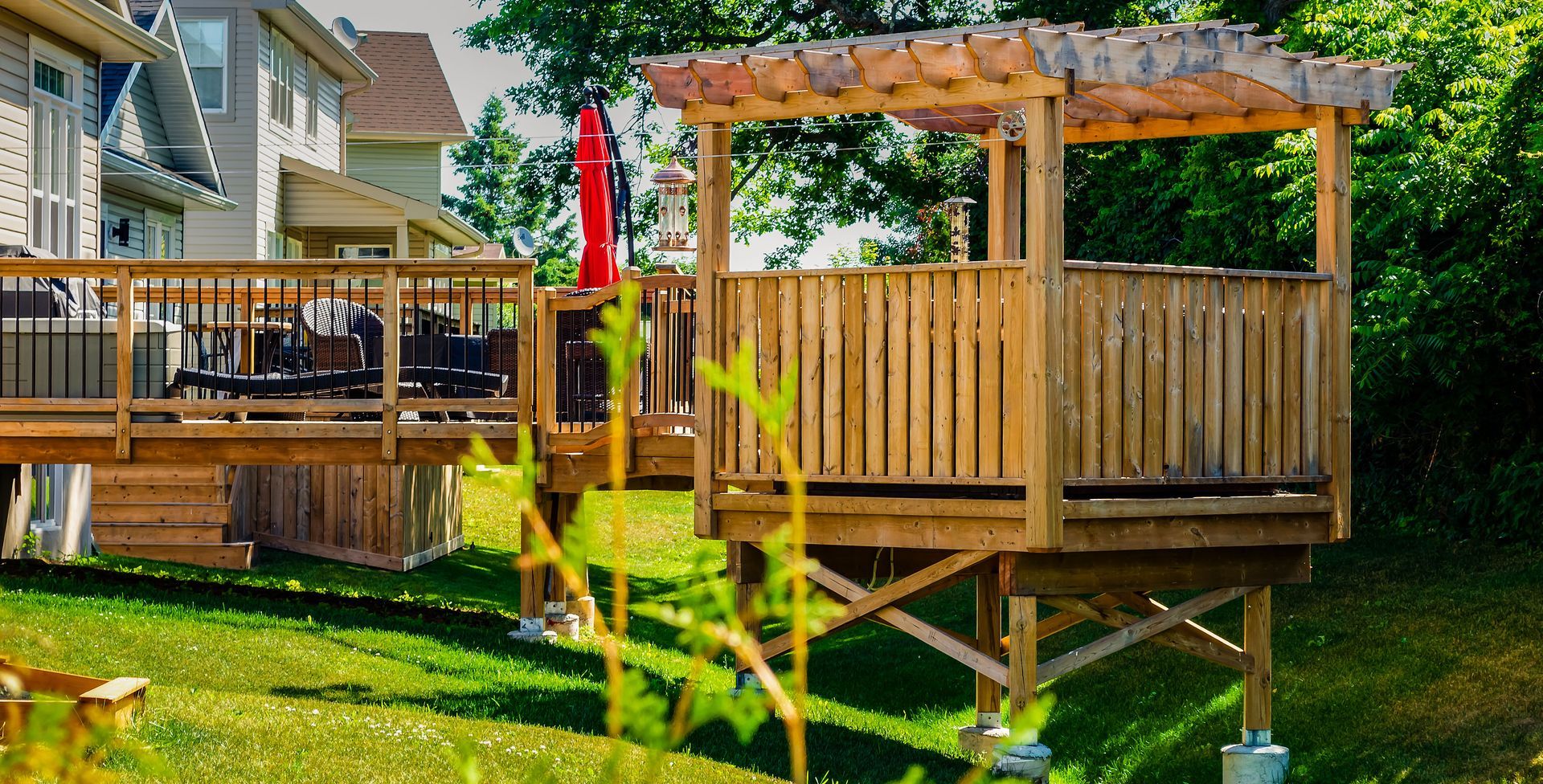 Wooden backyard deck with pergola, railings, and a red umbrella beside a house and green lawn