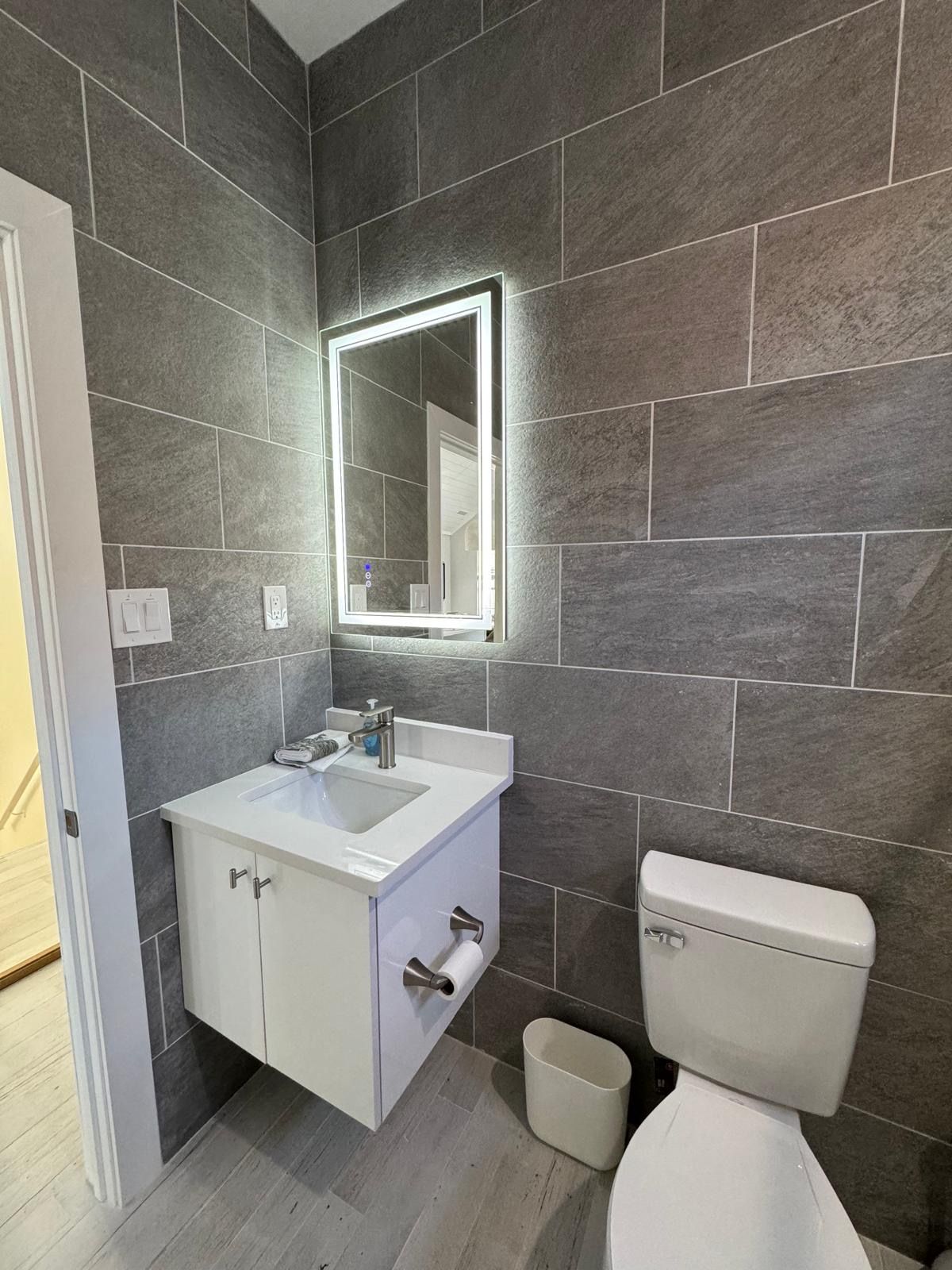 Modern gray-tiled bathroom with wall-mounted sink, lit mirror, and toilet