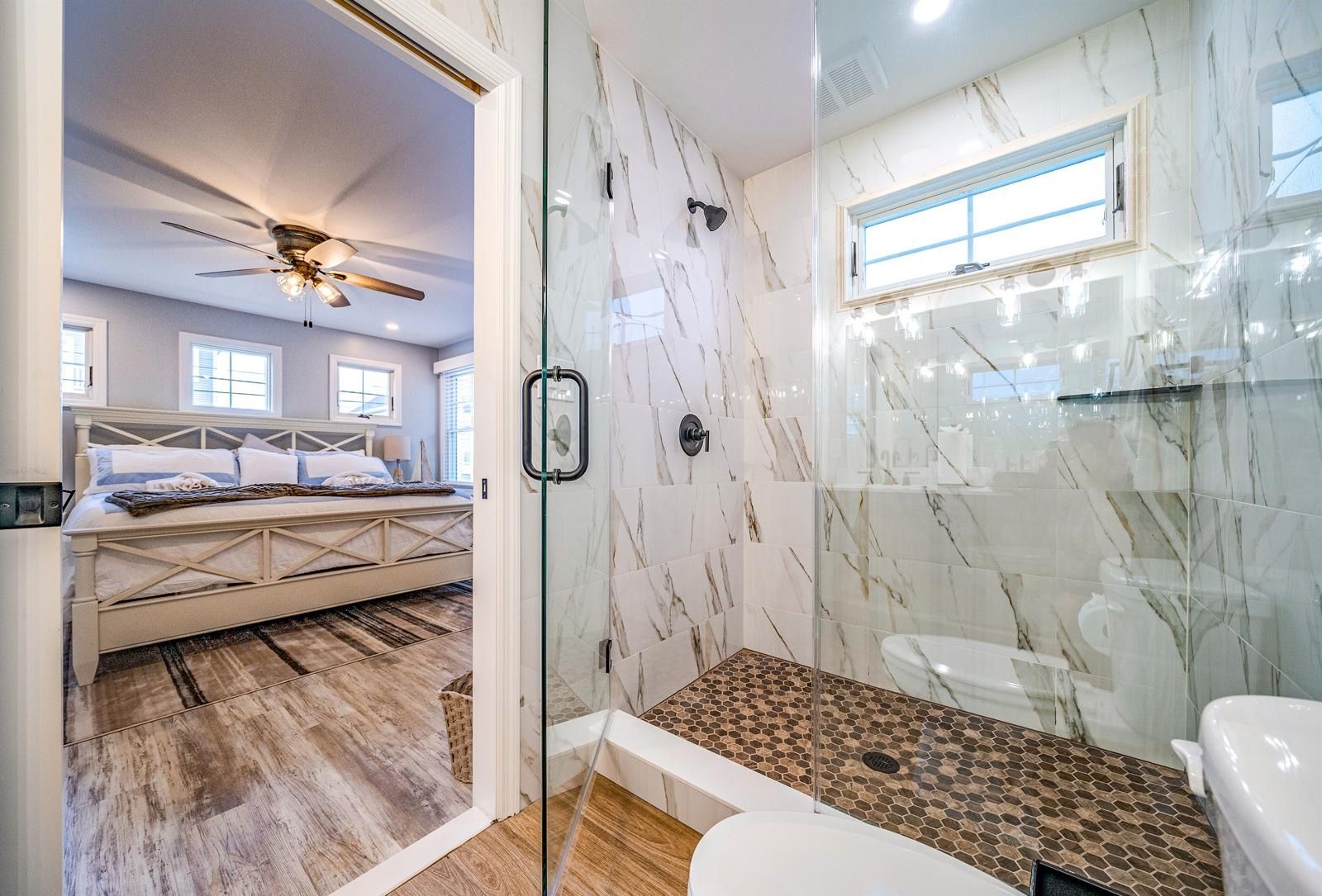 Bathroom with marble-tile shower beside a bedroom with ceiling fan and bed visible through the doorway