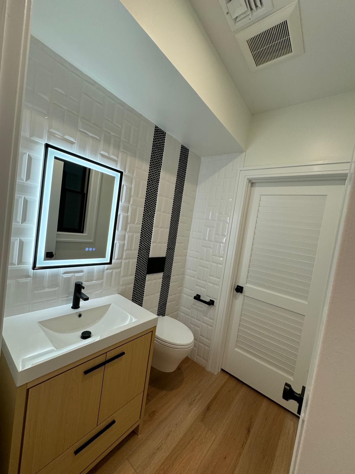 Modern bathroom with white vanity, mirror, toilet, black tile accent, and wood floor
