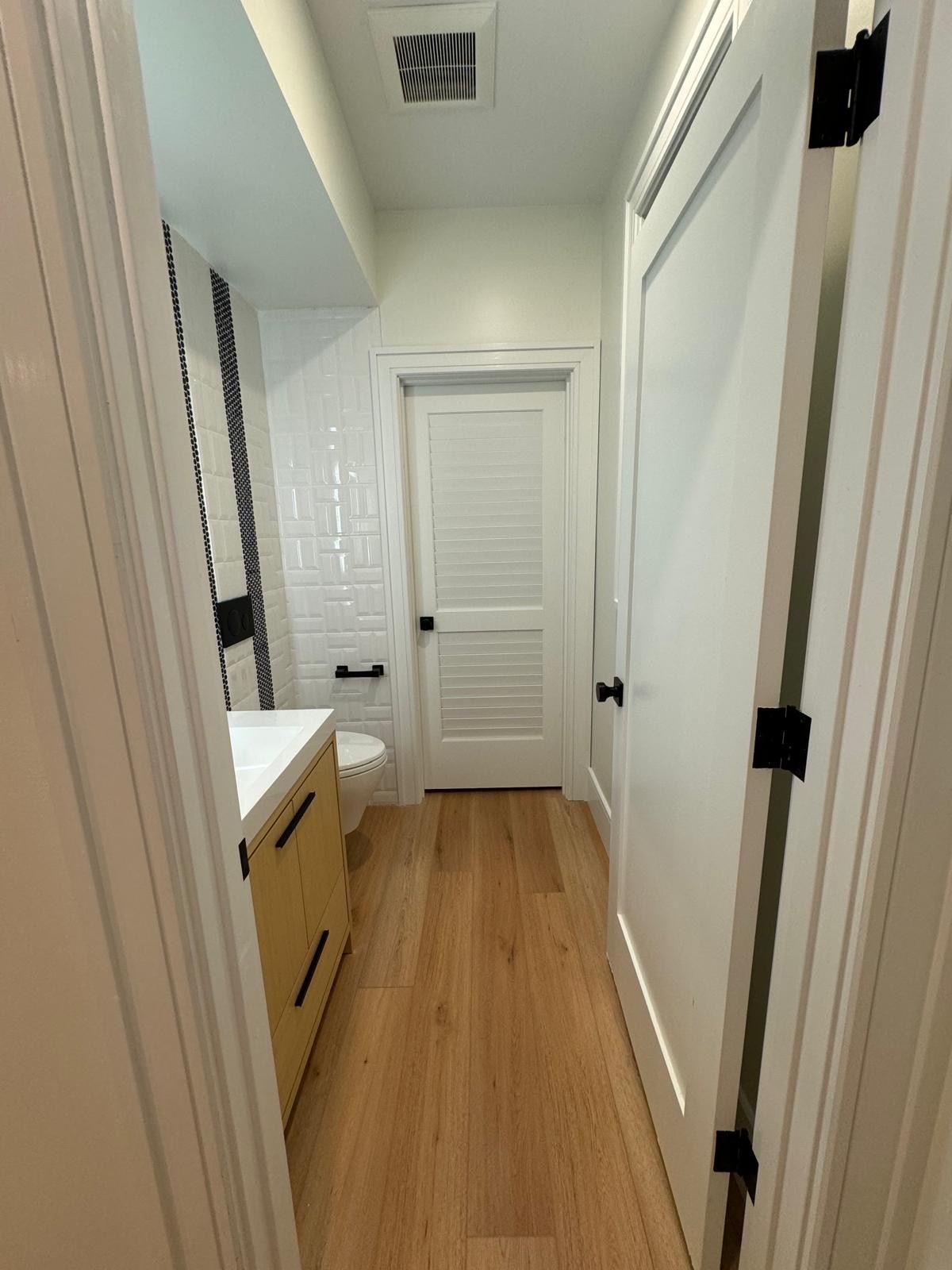 Narrow white bathroom with wood floor, vanity on left, closed door ahead, and wall vent above.