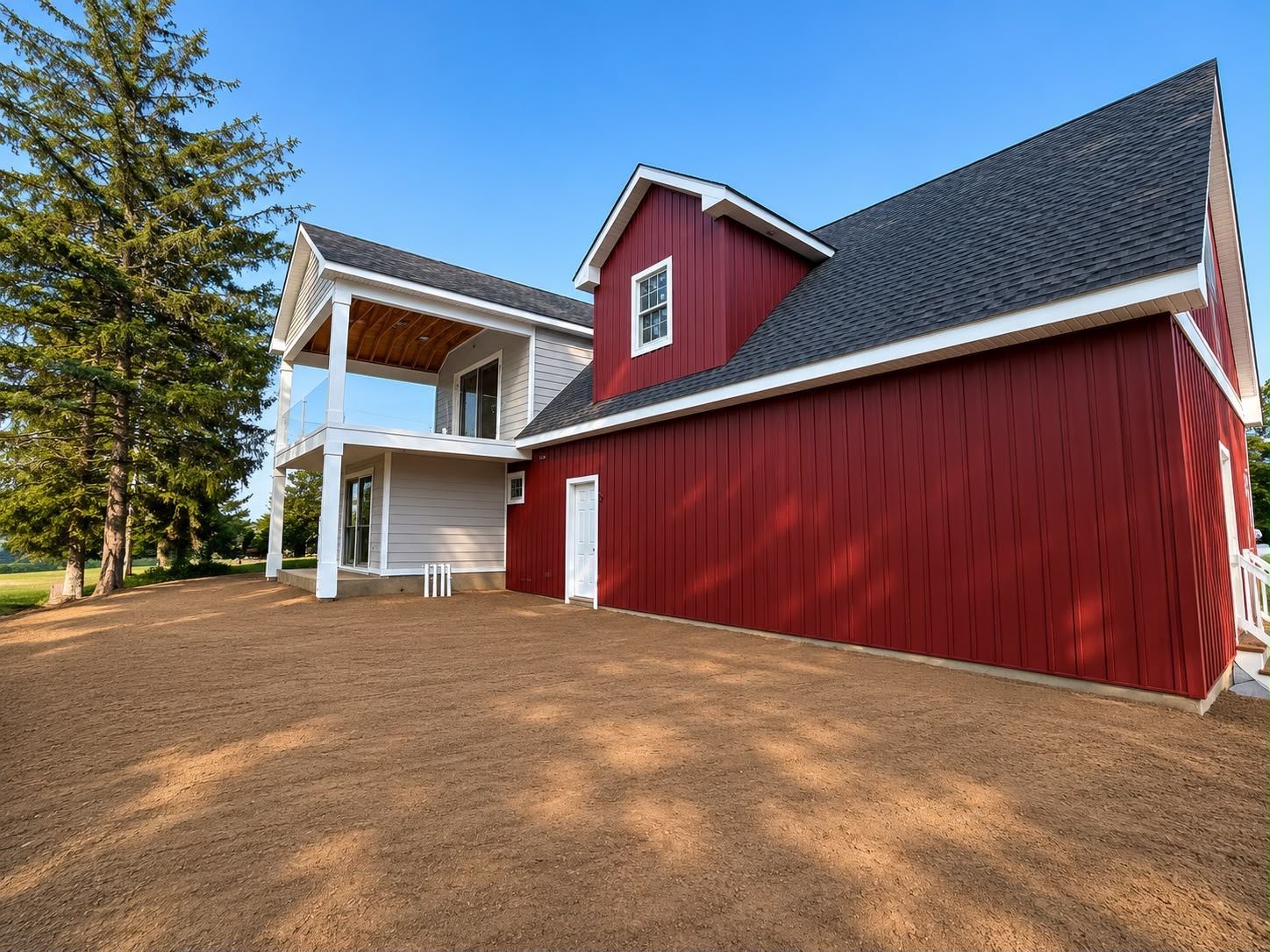 Red barn-style house with white porch on a dirt lot, viewed from the side under a clear sky