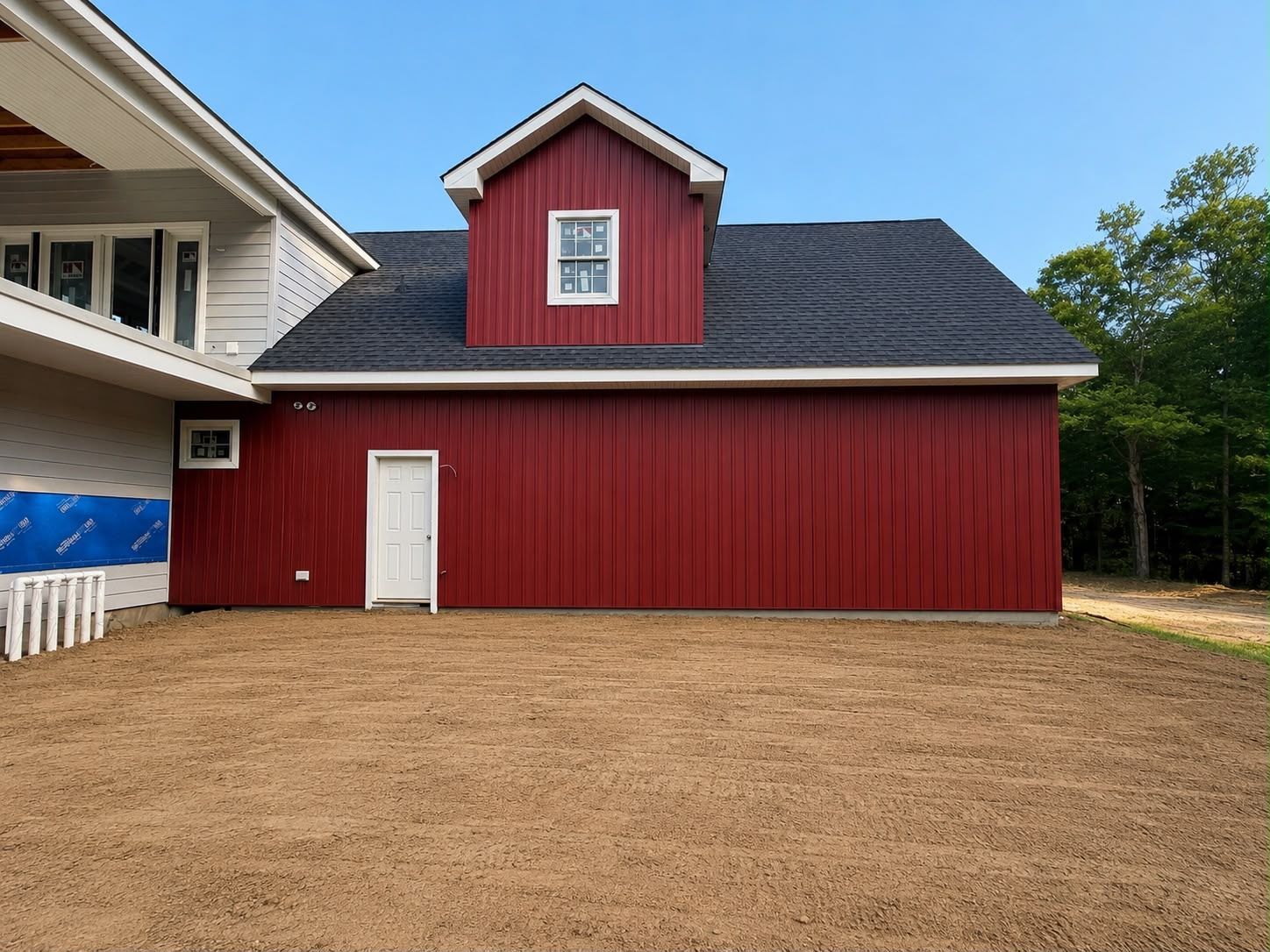 Red barn-style building with a white door and small upper window beside a house on a gravel lot.