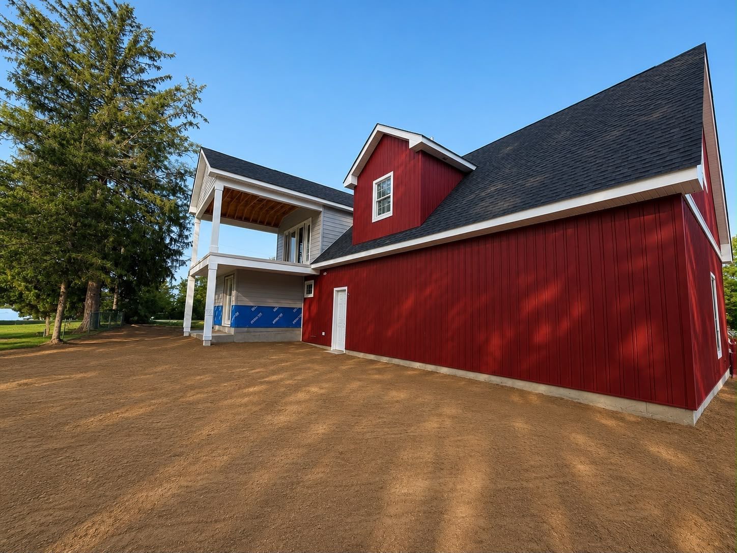 Red barn with white trim and a covered porch beside a tree on a gravel lot under a clear blue sky