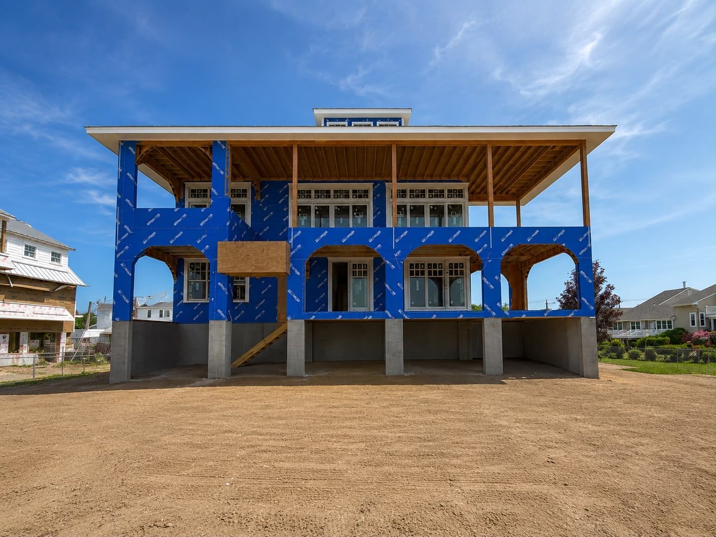 Blue two-story building under construction with open framing and a large dirt foreground under a clear sky