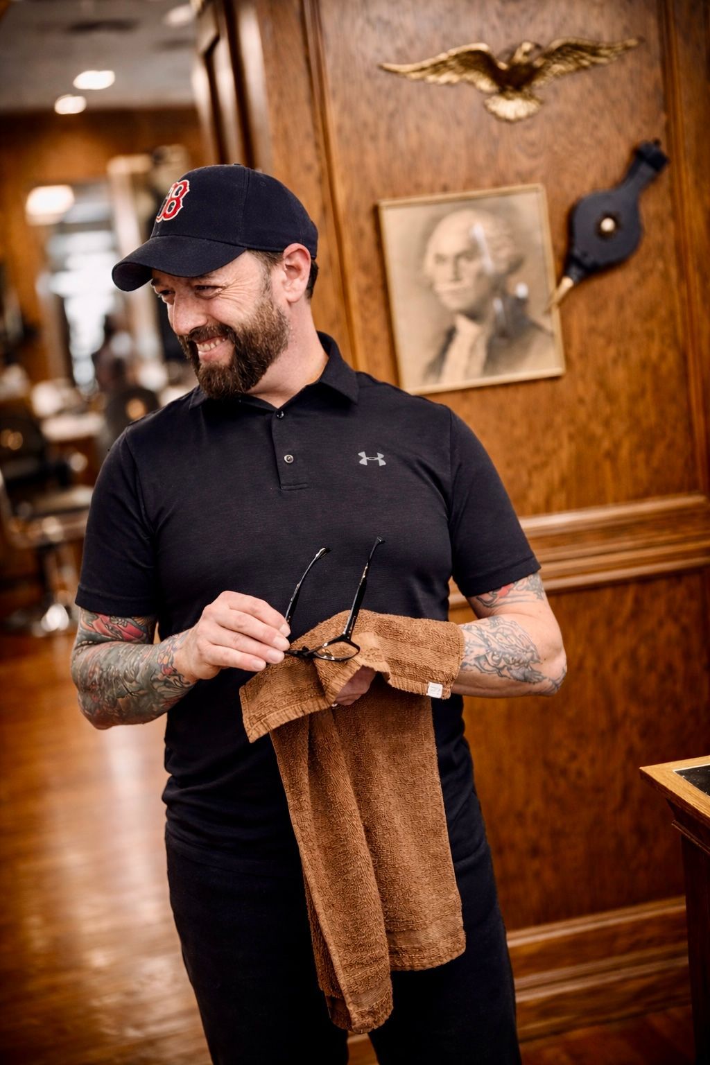 A smiling individual in a baseball cap and black polo wipes sunglasses with a towel inside a wood-paneled room.