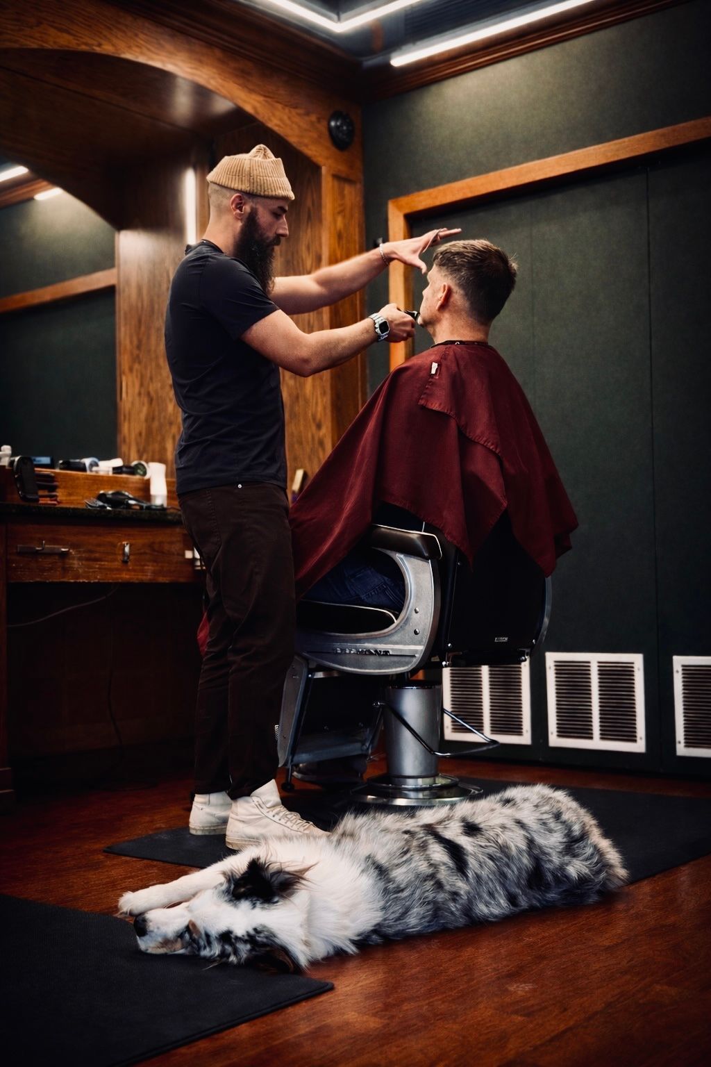 A barber cuts a customer's hair in a wood-paneled shop while a spotted dog sleeps on the floor.