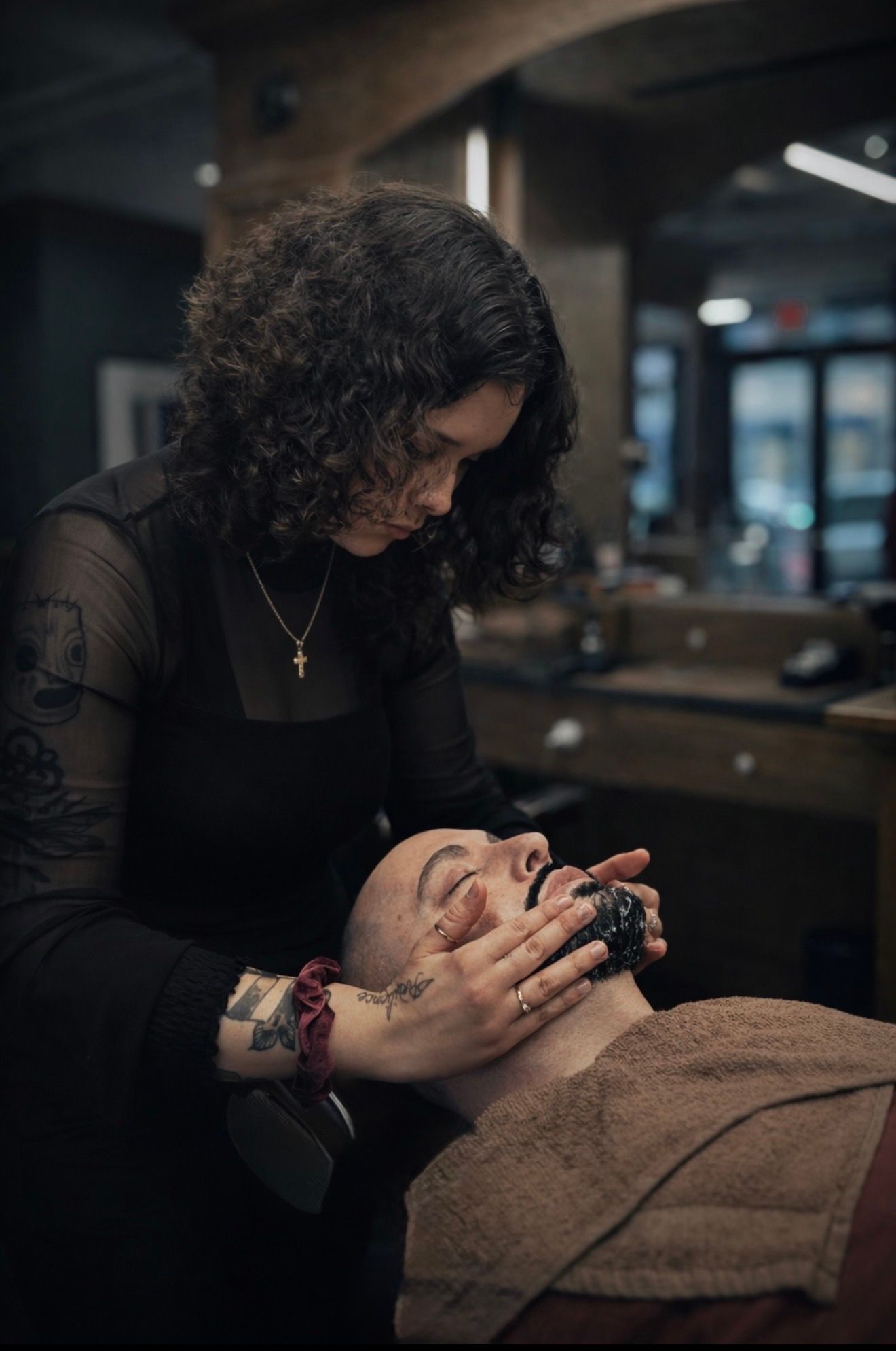 A barber in a black top applies hot lather to a client's face with a brush inside a dimly lit barbershop.