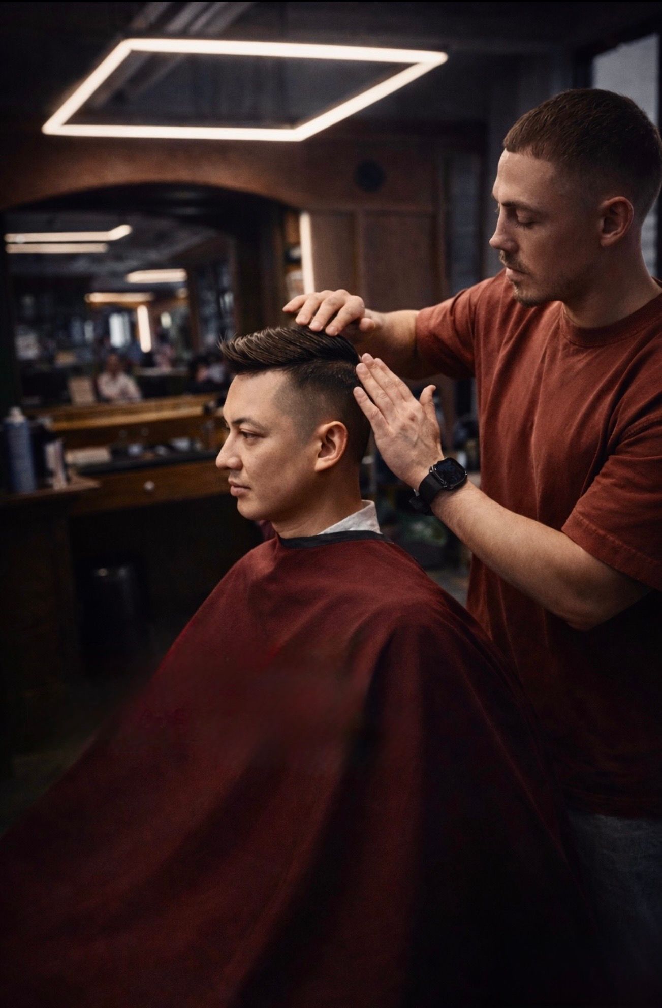 A barber in a rust-colored t-shirt styles a customer's hair in a dimly lit, modern salon.