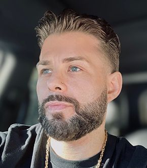 Man with short, light brown hair, full beard, wearing gold chain, looking off-camera.