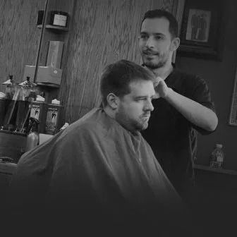 Barber cutting a customer's hair in a shop, both in black and white. The barber is focused, and the customer is looking down.