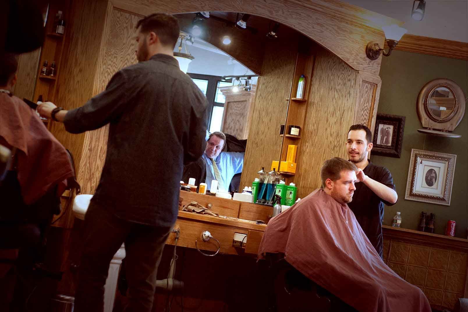 Barbers cutting hair in a shop, multiple mirrors, wood paneling, customers in capes.