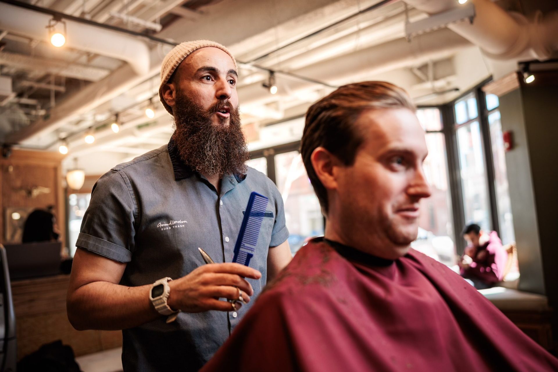 Barber giving a haircut. Man with beard and cap holding comb and scissors, client in red cape. Well-lit interior.