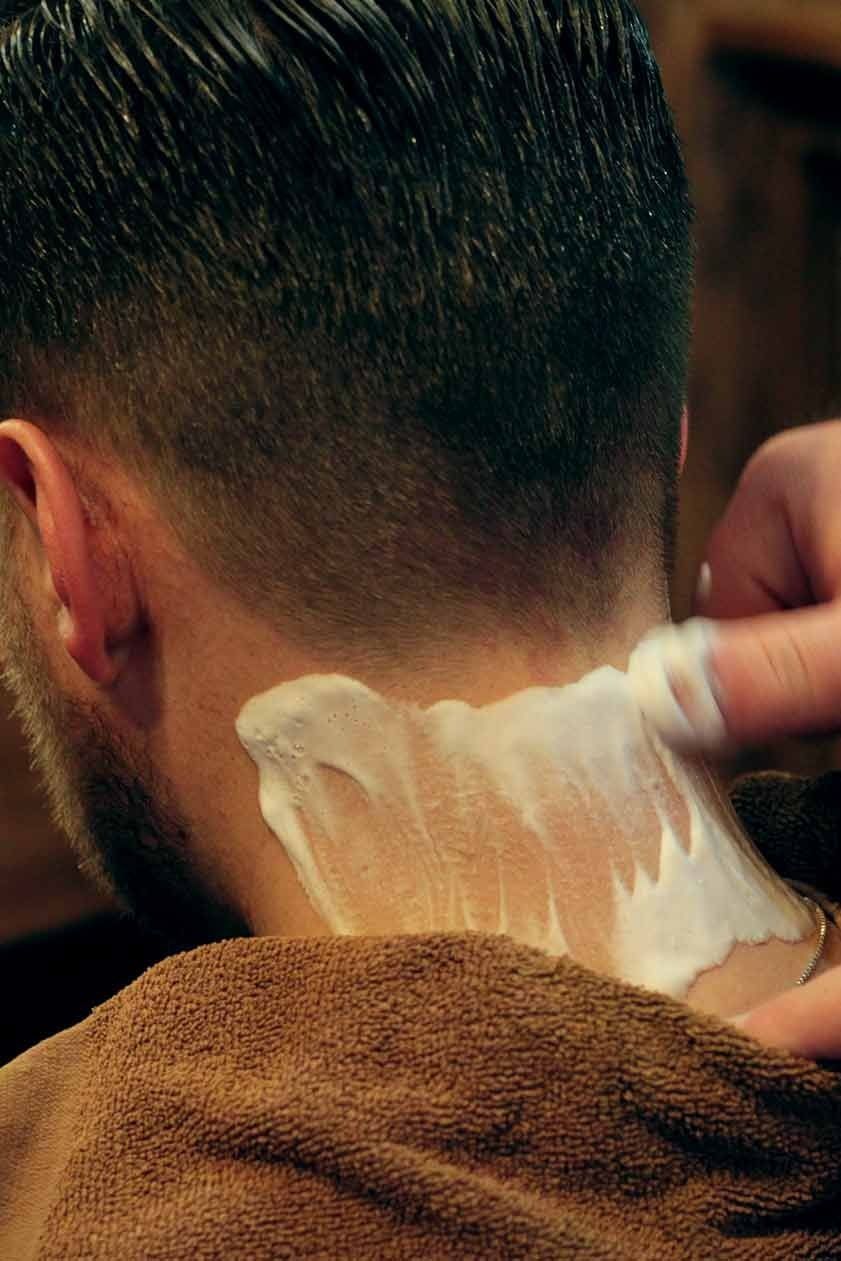 A barber shaving the back of a customer's neck, applying shaving cream with a hand.