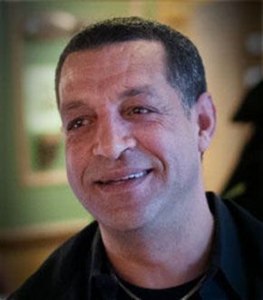 Man smiling, looking to the left. He has short dark hair, a black shirt, and appears to be indoors.