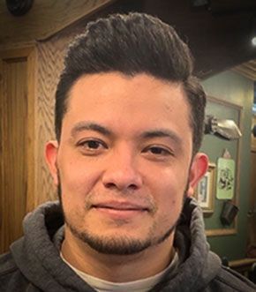 Man with dark hair and stubble in a gray hoodie smiles, indoors.