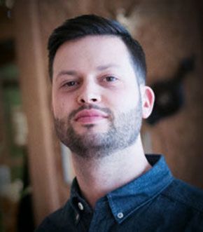Man with short dark hair and beard, wearing a blue button-down shirt, looking directly at the camera.