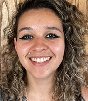 Woman with curly hair and a nose piercing smiles, wearing eyeliner.