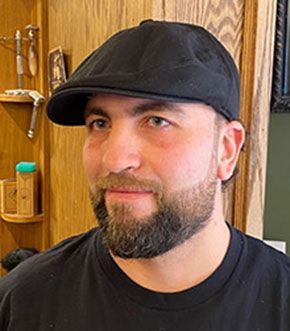 Man wearing a black cap and shirt, with a short beard, in front of a wooden wall.