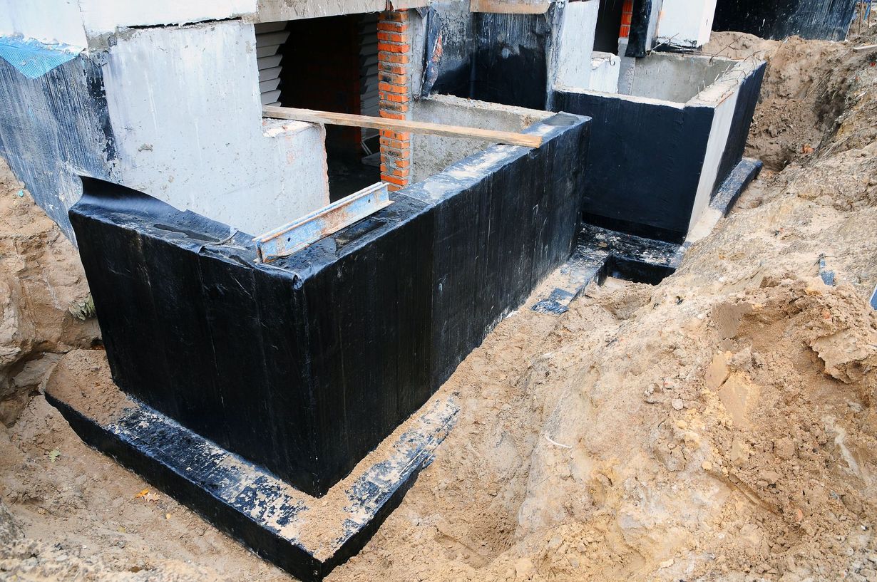 Black waterproofed concrete foundation in an excavation trench with exposed rebar and soil around it