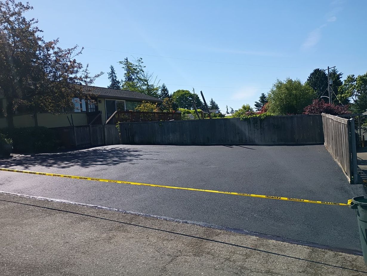 Fresh black asphalt driveway beside a brick house, with a car parked at left and garden edge at right