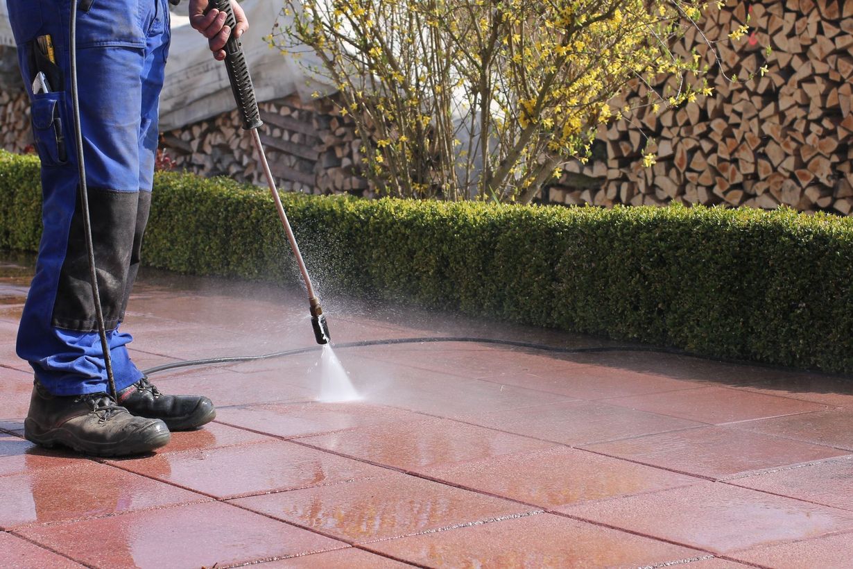 Person pressure-washing a red patio with a long wand near stacked firewood and hedges