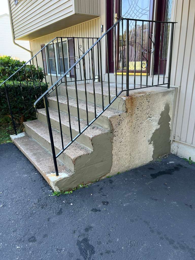 Concrete apartment steps with a black metal railing and chipped side wall beside asphalt pavement